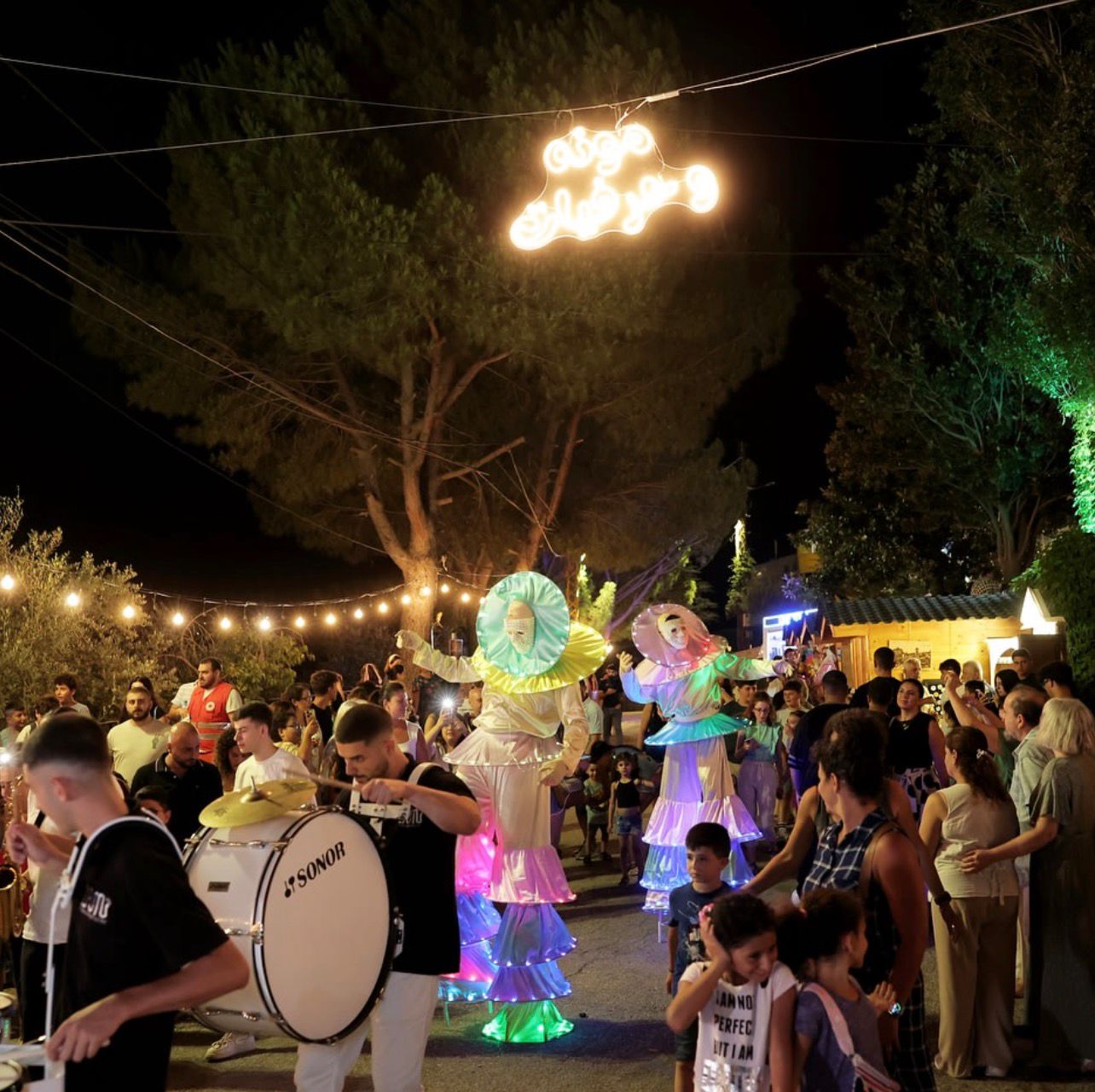 Night parade with illuminated performers, musicians, and families walking through a festive street in Kobayat.