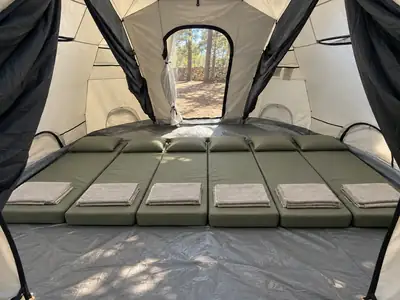 Inside a family camping tent with six mattresses made up with folded towels, open to the pine forest