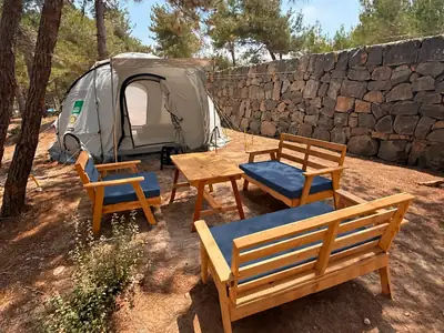 Dome tent with two cushioned benches, an armchair and a wooden table beside a stone retaining wall